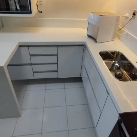 Cozinha com bancada branca em L, cuba de inox e armários brancos